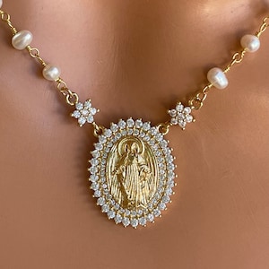 May include: A gold-tone necklace featuring a central oval pendant with a relief of a figure surrounded by clear crystals. The pendant is flanked by two small crystal-encrusted flower motifs, and the chain is accented with small white pearls.