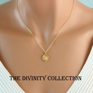 May include: A gold chain necklace with a small round pendant featuring a detailed design. The necklace is worn by a model with a white shirt. The text "THE DIVINITY COLLECTION" is visible at the bottom of the image.