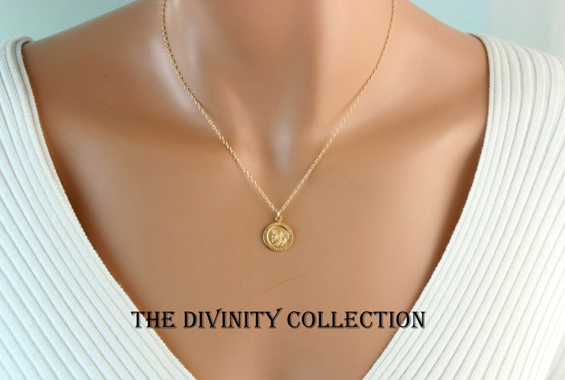 May include: A gold chain necklace with a small round pendant featuring a detailed design. The necklace is worn by a model with a white shirt. The text "THE DIVINITY COLLECTION" is visible at the bottom of the image.