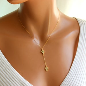 May include: Gold necklace with a hamsa hand charm and an evil eye charm. The necklace is on a mannequin wearing a white sweater.