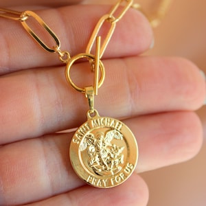 May include: Gold chain necklace with a gold pendant featuring an image of Saint Michael the Archangel and the text "Saint Michael Pray For Us".