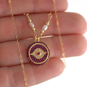 May include: Gold chain necklace with a pink evil eye pendant. The pendant is decorated with small clear and pink stones.