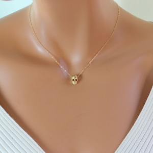 May include: A delicate gold-tone necklace featuring a small, detailed skull pendant suspended from a thin, matching link chain. The skull charm is centered on the chain, which rests against the collarbone area, providing a subtle and polished look.