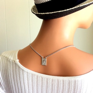 May include: A silver-toned necklace with a rectangular pendant featuring a religious image. The pendant hangs from a delicate chain. The background includes a white ribbed shirt and a straw hat with a black ribbon.