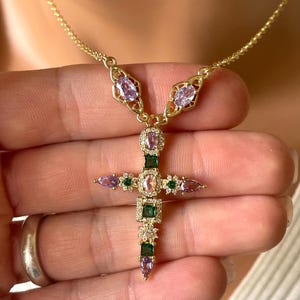 May include: A gold-tone necklace featuring a cross pendant adorned with oval and square-cut purple and green gemstones, accented by small clear crystals. The pendant is suspended from a delicate gold-tone chain with two additional oval purple stones.