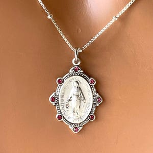 May include: Silver-toned pendant necklace featuring an oval pendant with an image of a religious figure. The pendant is adorned with small, round, red gemstones. The necklace has a delicate chain.