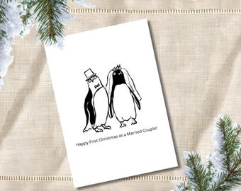 May include: A white greeting card featuring a black and white line drawing of two penguins, one wearing a top hat and bowtie, the other wearing a veil. Below the illustration, the text reads: Happy First Christmas as a Married Couple!