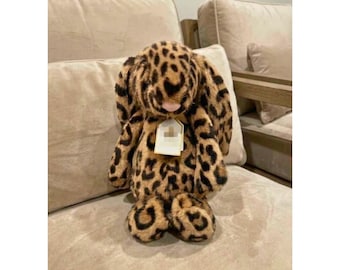 Peut inclure: Une peluche en forme de lapin avec un motif léopard marron et noir, de longues oreilles tombantes et un petit nez rose. Une étiquette rectangulaire blanche est attachée au centre de la poitrine, affichant un petit logo et du texte.