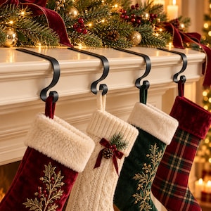 May include: Four black metal stocking holders with a curved hook design, each supporting a Christmas stocking. The stockings feature varied textures and patterns, including red velvet with gold embroidery, cream cable knit, green velvet, and red plaid.