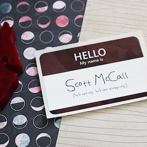 May include: A white name tag with a brown border. The text on the tag reads "HELLO My name is Scott McCall (We can try. We can always try.)"