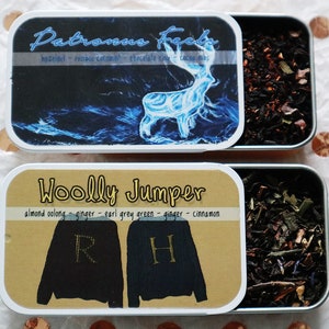 May include: Two open metal tins of loose leaf tea. One tin has a label that reads "Patronus Feels" with a blue deer illustration. The other tin has a label that reads "Woolly Jumper" with sweater illustrations. The tea leaves are visible.