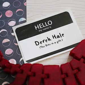 May include: A black and white name tag with the text "HELLO my name is Derek Hale (The Bite is a gift.)" on a white background. The name tag is on a wooden surface with a red plastic toy in the foreground.
