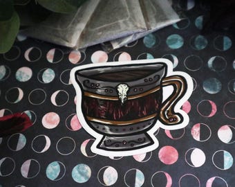 Op de afbeelding: Een sticker van een donkergrijze keramische mok met een gouden rand en een schedelhandvat. De mok is gevuld met een donkerbruine vloeistof en heeft een rood en gouden ontwerp aan de zijkant.