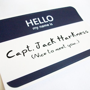 May include: A white name tag with a navy blue top section. The text "HELLO my name is" is printed in white. Below, handwritten text reads "Capt. Jack Harkness (Nice to meet you...)".