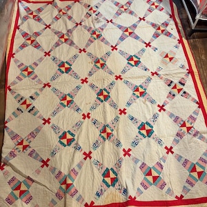 May include: A rectangular quilt featuring a repeating star pattern in various floral and geometric prints. The design includes off-white fabric sections, red cross-shaped accents at the intersections, and a red border with a thin yellow trim.
