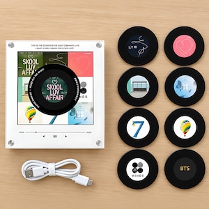 May include: A square white acrylic music player display frame featuring a central black circular disc, accompanied by eight interchangeable black circular magnetic discs with various graphic designs and a white USB charging cable.