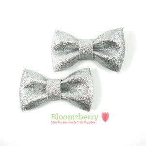 May include: Two silver glitter bow hair clips. The bows are made of fabric and have a shiny, sparkly finish.