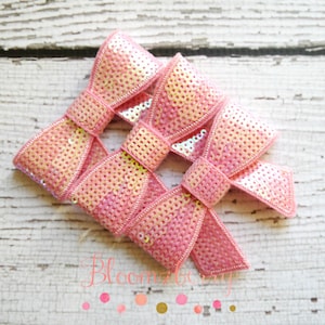 May include: Three pink sequined bows with a shimmery finish. The bows are arranged in a cluster on a white wooden surface.