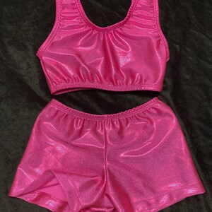 Op de afbeelding: Een roze, glinsterende turnpak en shorts set voor gymnastiek of dans. Het topje is mouwloos en heeft een ronde hals. De shorts zijn kort en hebben een brede tailleband.
