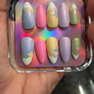 May include: A set of ten almond-shaped press-on nails in a clear plastic case. The nails feature a variety of pastel colors including blue, pink, yellow, lavender, and green, with small raised mouse-ear shaped accents on several nails.