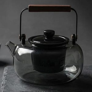 May include: A clear, smoke-tinted glass teapot featuring a black ceramic lid and a matching black internal strainer. The teapot has a black metal handle with a dark brown wooden grip, a short spout, and a rounded, wide-bottomed glass body.
