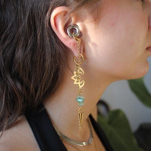 Op de afbeelding: Een goudkleurig oorgewicht met een ronde metalen plug, een bungelende ketting, een lotusbloem-bedel, kleine lichtgroene stenen kralen en een puntige goudkleurige stekel. Het sieraad hangt aan de oorlel en toont een gelaagd metalen ontwerp.