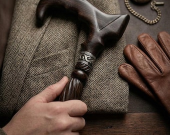 May include: A dark brown wooden walking cane handle with a curved grip and a silver-toned metal collar featuring intricate scrollwork engravings. The handle is positioned over a brown herringbone patterned blazer next to leather gloves.