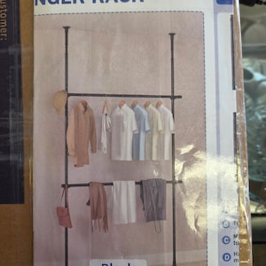 May include: A black floor-to-ceiling tension hanger rack with two horizontal bars for hanging clothes. The rack features vertical poles and holds various shirts, a scarf, a bag, and a hat. The text NGER RACK and Black are visible.