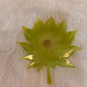 May include: A leaf-shaped resin tray in a translucent lime green colour with a smooth, glossy finish. The centre of the tray features a circular cluster of fine gold glitter, and the edges are pointed and jagged, mimicking a natural maple leaf.