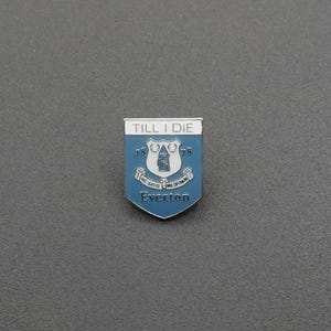 以下が含まれることがあります： 盾の形をした青と白のエナメルピン。上部には「TILL I DIE」の文字が刻まれています。中央には塔のグラフィックがあり、両側に数字の18と78が配置されています。その下にはラテン語のテキストと「Everton」という単語が書かれたバナーがあります。