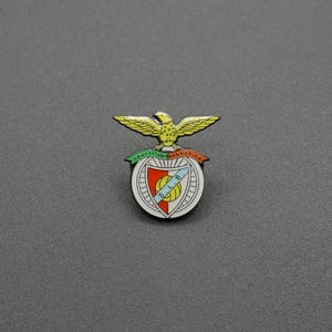 以下が含まれることがあります： Sport Lisboa e Benficaのエンブレムが描かれた金属製のラペルピン。黄色い鷲が赤、白、緑のアクセントが入った円形の盾の上に止まっており、中央の青い斜めのストライプには金色の文字でS.L.B.と記されています。