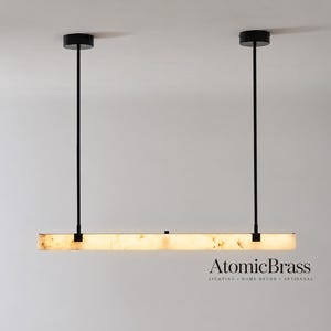 May include: A linear pendant light fixture featuring a long, rectangular, translucent stone bar with natural veining, suspended by two thin black metal rods from circular black ceiling mounts. The text AtomicBrass is visible.