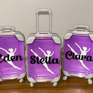 May include: Three small, purple rolling suitcases with white and silver accents. Each suitcase has a white silhouette of a dancer and a name in black script: Eden, Stella, and Clara. The suitcases have a handle that can be extended.