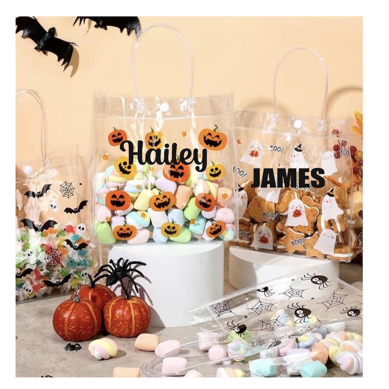 May include: Clear plastic Halloween treat bags filled with candies and cookies. One bag has the name "Hailey" with pumpkin graphics, and another says "JAMES". Decorative pumpkins and spiders are also present.