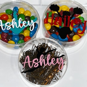 May include: Three clear plastic containers with lids. Each container has a different design and name. The first container has a blue and white cheerleader design with the name "Ashley" and is filled with colorful M&Ms. The second container has a black and red cheerleader design with the name "Hailey" and is filled with colorful M&Ms. The third container has a black and pink dancer design with the name "Ashley" and is filled with brown bobby pins.