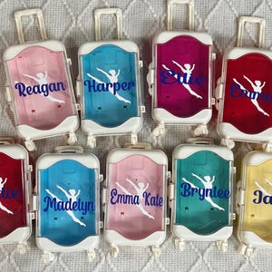 May include: A collection of miniature, personalized rolling suitcases in various colors, including pink, blue, red, and yellow. Each suitcase features a name in blue script and a white dancer silhouette.