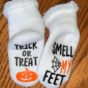 K&ouml;nnte beinhalten: Ein Paar wei&szlig;e Halloween-Socken. Auf einer Socke steht "TRICK OR TREAT" mit einem orangefarbenen K&uuml;rbis. Auf der anderen steht "SMELL MY FEET" mit einem schwarzen Spinnennetz und orangefarbenem Text.