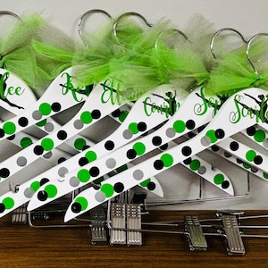 May include: White wooden hangers with green, black, and gray polka dots. Each hanger has a name in green script and a green tulle bow. The hangers are designed for dance or gymnastics apparel.