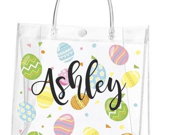Puede incluir: Bolso tote transparente con asa transparente y cierre de botón a presión. La bolsa está decorada con coloridos huevos de Pascua y el nombre "Ashley" en escritura negra. Los huevos son de varios colores y patrones.