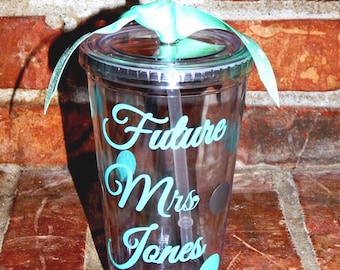 May include: Clear plastic tumbler with a lid and straw. The tumbler has the words "Future Mrs Jones" in teal script. Teal polka dots and a teal ribbon adorn the cup. The cup is sitting on a brick surface.