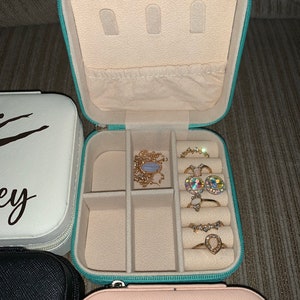 May include: An open, turquoise jewelry travel case with a cream interior, containing rings and a necklace. The case is zipped and has storage compartments. Other closed jewelry cases are visible.