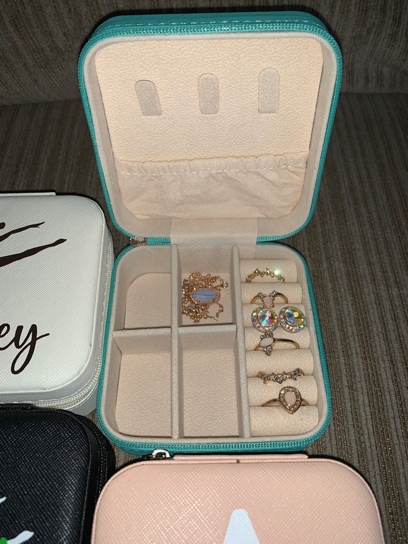 May include: An open, turquoise jewelry travel case with a cream interior, containing rings and a necklace. The case is zipped and has storage compartments. Other closed jewelry cases are visible.
