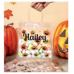 May include: Clear plastic Halloween-themed gift bag filled with colorful marshmallows. The bag is decorated with jack-o'-lantern stickers and the name "Hailey" in black script. Two orange pumpkins are in the background.