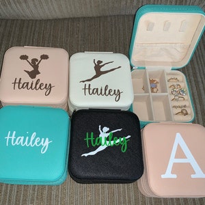May include: Several small, square jewelry cases in shades of teal, black, and pink. Each case is personalised with a name or initial and a silhouette of a dancer or cheerleader. One case is open, showing compartments for jewellery.