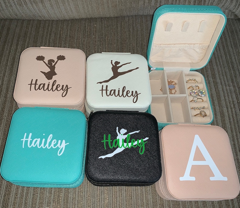 May include: Several small, square jewelry cases in shades of teal, black, and pink. Each case is personalised with a name or initial and a silhouette of a dancer or cheerleader. One case is open, showing compartments for jewellery.