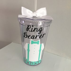 May include: Clear plastic tumbler with a white bow on top. The tumbler has a black vinyl decal that says "Ring Bearer" with a cartoon image of a bow tie, white shirt, and green suspenders. The name "Chase" is written in green below the image.