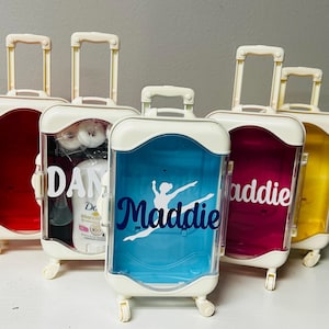 May include: Five small, white-framed rolling suitcases with clear, colored fronts. The suitcases have white handles and wheels. One suitcase has a red front, one has a blue front with a dancer graphic and the name "Maddie", one has a pink front with the name "Addie", and one has a yellow front.