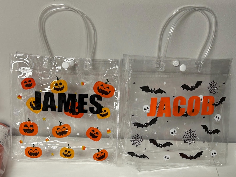 May include: Clear plastic Halloween treat bags with clear handles. One bag has orange pumpkins and the name "JAMES" in black. The other bag has black bats, spiderwebs, skulls, and the name "JACOB" in orange.