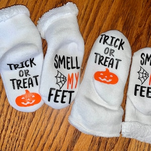 K&ouml;nnte beinhalten: Vier Paar wei&szlig;e Halloween-Socken. Jede Socke zeigt die S&auml;tze "Trick or Treat" und "Smell My Feet" in schwarzer Schrift, zusammen mit einem orangefarbenen K&uuml;rbis und einer Spinnennetz-Grafik. Die Socken haben einen weichen, flauschigen Bund.