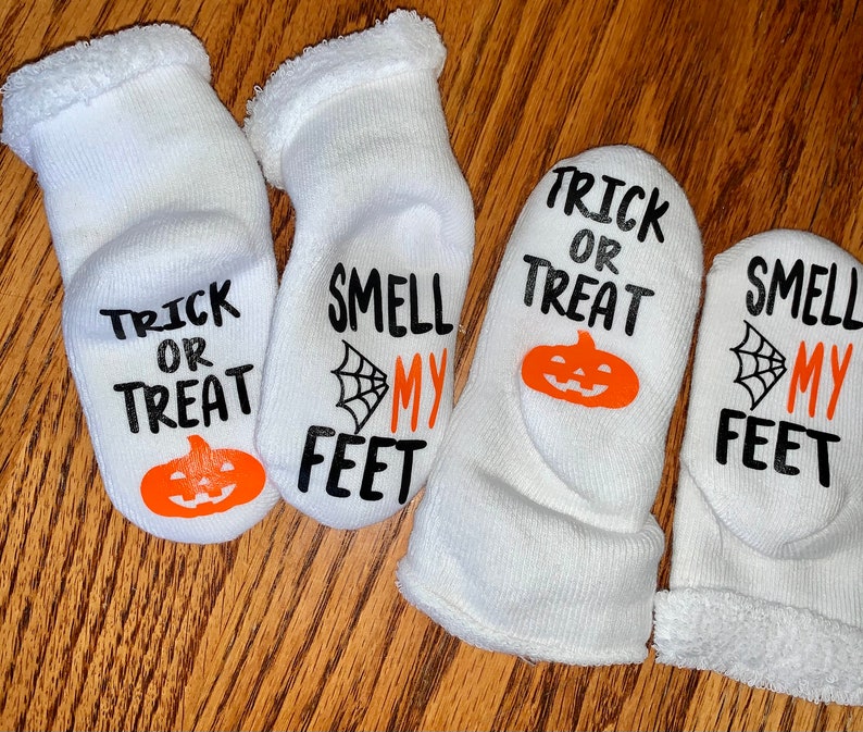K&ouml;nnte beinhalten: Vier Paar wei&szlig;e Halloween-Socken. Jede Socke zeigt die S&auml;tze "Trick or Treat" und "Smell My Feet" in schwarzer Schrift, zusammen mit einem orangefarbenen K&uuml;rbis und einer Spinnennetz-Grafik. Die Socken haben einen weichen, flauschigen Bund.